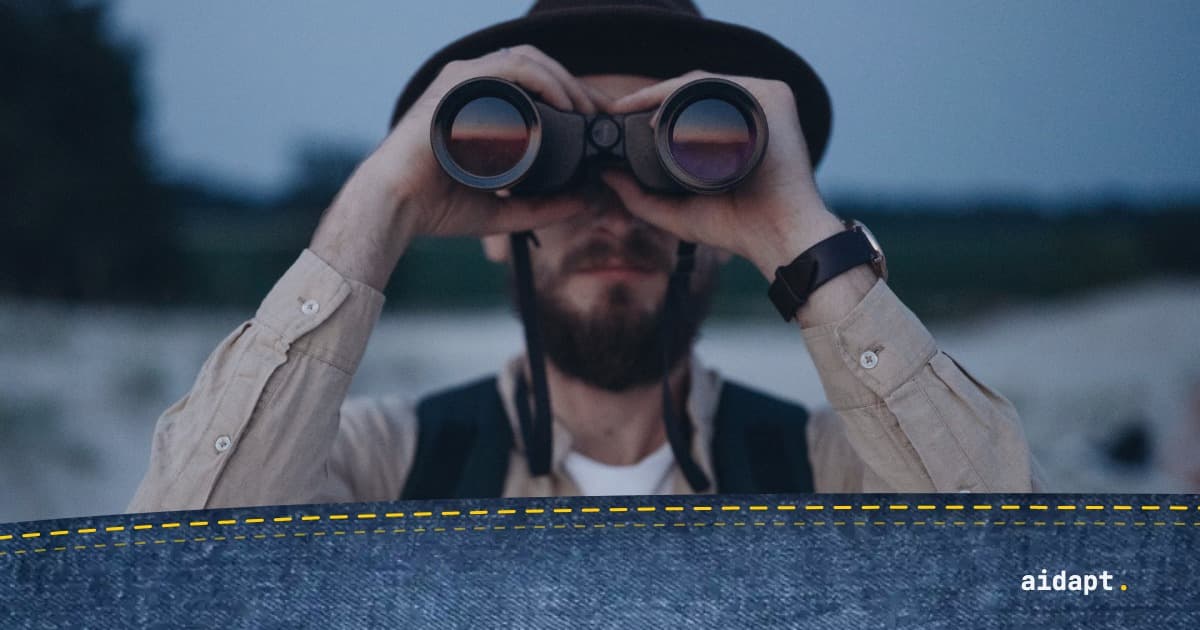 Man med hatt och skägg tittar genom en kikare i skymningen, sanddyner i bakgrunden