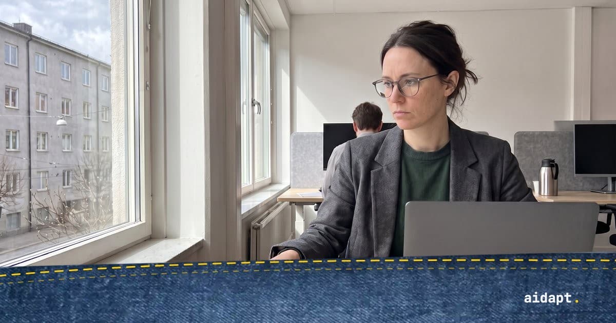 Kvinna i glasögon och grå kavaj läser på en laptop vid ett björkskrivbord i ett ljust kontor med höga fönster
